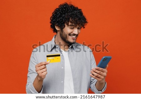 Smiling young bearded Indian man 20s years old wears blue shirt using mobile cell phone hold credit bank card doing online shopping order delivery isolated on plain orange background studio portrait Royalty-Free Stock Photo #2161647987