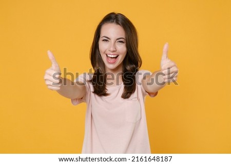 Cheerful funny laughing young brunette woman 20s wearing pastel pink casual t-shirt posing standing showing thumbs up looking camera isolated on bright yellow color wall background studio portrait