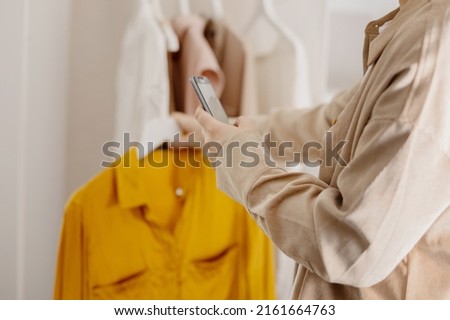 Woman holding smartphone and taking photo of her old clothes to sell them online. Selling on website, e-commerce. Reuse, second-hand concept. Conscious consumer, sustainable lifestyle. Close-up view.