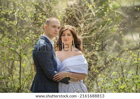a guy in a suit and a girl in a white dress are standing in the forest and hugging
