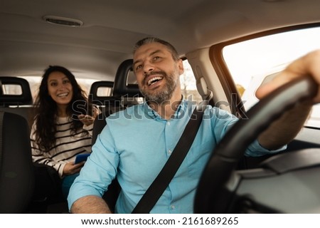 Nice Ride. Portrait of happy male driver riding car looking at cheerful beautiful lady sitting inside auto on back passenger seat, female using cell phone and talking with guy, windshield view Royalty-Free Stock Photo #2161689265