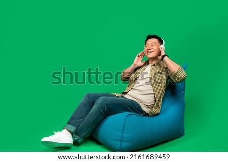Rest and relax. Mature asian man sitting on beanbag chair and listening to music with closed eyes, enjoying great sound in new wireless headphones, green background, free space