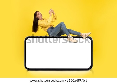 Cheerful Casual Lady Sitting On Big Giant Huge Horizontal Smartphone With Empty White Display, Excited Female Holding Using Cell Phone, Playing Games, Mock Up, Full Body Length Banner, Copy Space Royalty-Free Stock Photo #2161689753