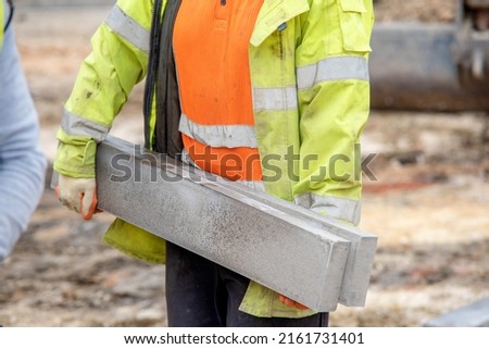 Close up of groundworker in orange and yellow hi-viz  carrying heavy concrete kerbs on construction site during new road construction Royalty-Free Stock Photo #2161731401