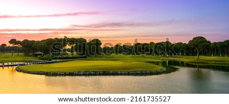 Panoramic view of beautiful golf course with pines at sunset. Golf field with fairway, green, hole, flag and lake