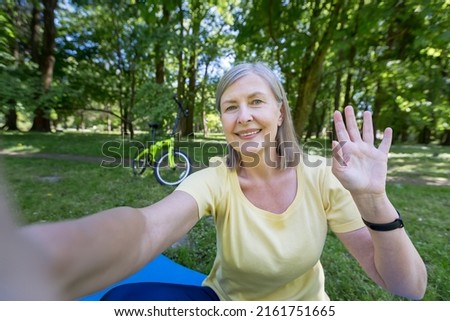Senior gray-haired woman, active sportswoman in the park in the summer looks at the phone camera and smiles waving her hand at a video call