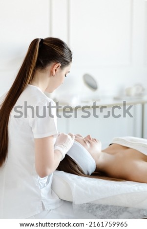 A female cosmetologist makes facial procedures for a client in an aesthetic medicine clinic. Cosmetologist or dermatologist doing peeling or lifting massage to woman in spa or salon.High quality photo Royalty-Free Stock Photo #2161759965
