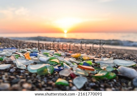 Colored sea glass with beach pebbles and shells in the mediterranean coast and in the background sea and waves with sunset and sky, sandy beach with coast glass, beach glass. Royalty-Free Stock Photo #2161770091