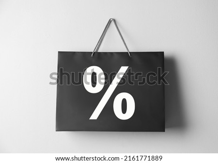 Black paper shopping bag with percent sign hanging on white wall. Discount concept