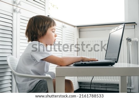 Boy sits at the table, uses laptop, typing on a keyboard, looks at the screen. Child does homework lesson, plays video game, study IT course. Home distance online education concept. Smart technology.
