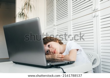 Boy lies at the table, looks at the computer screen. Tired pupil. Difficult, boring, uninterested and long study program. Child does homework lesson. Bad vision. Incorrect posture. Smart technology. Royalty-Free Stock Photo #2161788537