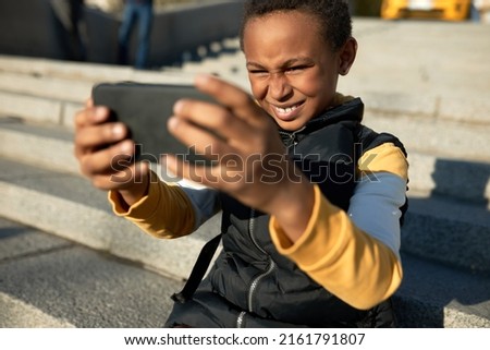 Selective focus of happy school boy with dark skin in urban style clothes having fun holding smartphone and making selfie, sitting on concrete stairs, skipping school, frowning in bright sunlight