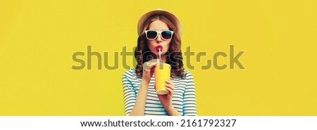 Portrait of stylish young woman drinking fresh juice wearing summer straw hat, sunglasses and striped t-shirt on yellow background Royalty-Free Stock Photo #2161792327