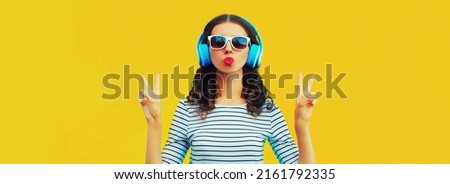 Portrait of stylish young woman in headphones listening to music blowing her lips sends sweet air kiss on yellow background, blank copy space for advertising text