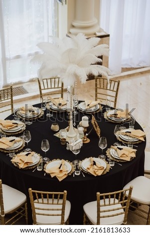 Wedding table setting in gatsby style with feathers. black tablecloths and gold plates. on a background of a white pompous hall top view Royalty-Free Stock Photo #2161813633