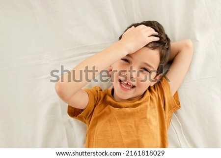 Portrait of cute cheerful child lying on the bed, looking at you. Happy seven year old brunette boy in a yellow 
t-shirt. Top view. Concept of rest, bed linen, happiness, face expression, son. Royalty-Free Stock Photo #2161818029