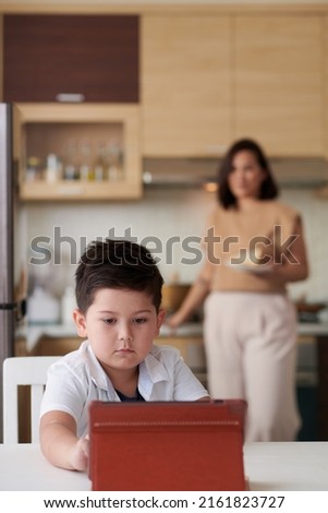 Cute little boy watching cartoons on tablet computer when mother cooking breakfast in background