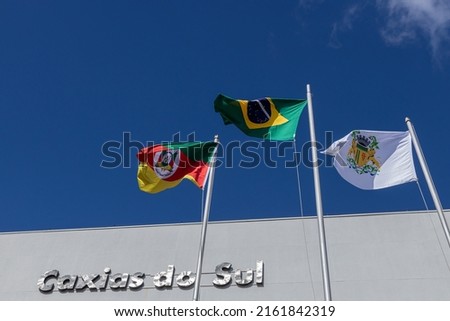 Brazil, Rio Grande do Sul state, and Caxias do Sul city flags flying in the wind on a sunny day. Blue sky background. Caxias do Sul sign on the wall