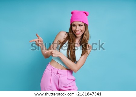 Attractive young woman in pink hat smiling and pointing away while standing against blue background