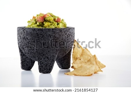 Traditional Mexican guacamole with avocado, onion, tomato, lime and green chili pepper in a typical mortar called molcajete by some totopos or nachos on a white background. Royalty-Free Stock Photo #2161854127