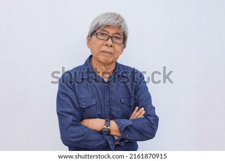 Asian senior grey-haired man in denim casual style with arms crossed  looks directly at camera with confident expression wears transparent eyeglasses isolated on white background.