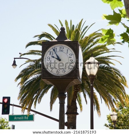 Large 4 sided Clock tower on tall pole with palm tree and traffic light blue sky background	
