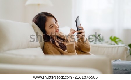 Happy young asian woman relax on comfortable couch at home texting messaging on smartphone, smiling girl use cellphone, browse wireless internet on gadget, shopping online from home Royalty-Free Stock Photo #2161876605