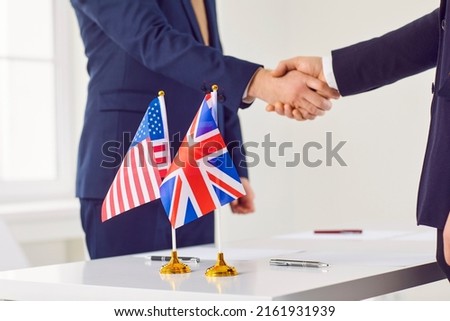 Diplomats from Britain and America meet together in office, reach bilateral trade agreement, sign business contract and exchange handshakes over negotiation table with national flags of USA and UK