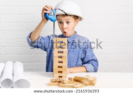 Little boy builder engineer in white construction helmet plays and builds high skyscraper from wooden blocks or cubes and plans. Emotional surprised child measures the tower with a measuring tape. Royalty-Free Stock Photo #2161936393