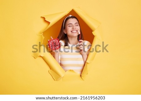 Beautiful satisfied woman wearing striped shirt and hair band, breaks through yellow paper background, holding alarm clock and toothbrush, wake up in good mood, smiling. Royalty-Free Stock Photo #2161956203