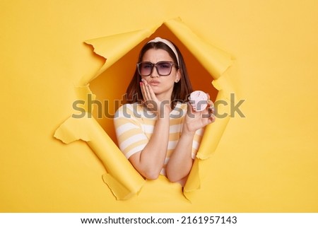 Portrait of sad unhealthy woman wearing striped shirt and hair band, breaks through yellow paper background, eating sweets and feels pain in tooth, suffering toothache.