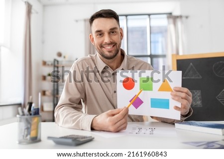 distance education, primary school and remote job concept - happy smiling male teacher with picture of geometric shapes in different colors having online class at home office