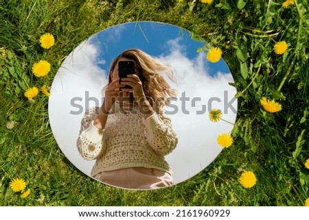 nature and people concept - woman with smartphone reflection in round mirror on summer field with dandelion flowers
