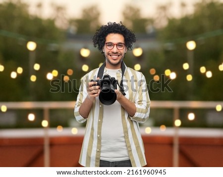 photography, profession and people and concept - happy smiling man or photographer in glasses with digital camera over party lights on roof top background
