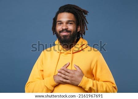 Calm young man of African American ethnicity 20s he has dreadlocks wearing casual yellow streetwear hoodie put folded hands on heart isolated on plain royal dark blue wall background studio portrait