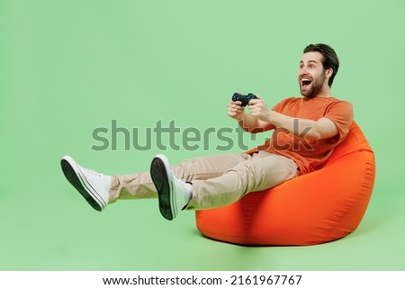 Full body young man wear casual orange t-shirt sit in bag chair hold in hand play pc game with joystick console isolated on plain pastel light green color background studio. People lifestyle concept
