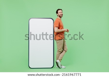 Full body side view young man wear casual orange t-shirt stand near mobile cell phone with blank screen workspace area isolated on plain pastel light green color background. People lifestyle concept