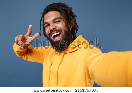 Close up funny man of African American ethnicity 20s he has dreadlocks wearing casual yellow hoodie doing selfie shot pov on mobile cell phone isolated on plain royal dark blue wall background studio