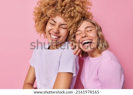 Happy pretty women laugh positively express happy sincere emotions have friendly relationship spend free time together stands closely dressed in casual t shirt isolated over pink background.