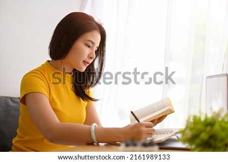 Charming Asian female university student sits in the library, research some informations on the book and taking notes on her school notebook.