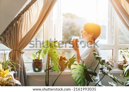 Side view woman drinking tea and looking at the sunrise or sunset while standing at the window in a room with green house plants, enjoying the moment. Relaxing and self-care, personal fulfillment. Royalty-Free Stock Photo #2161993035