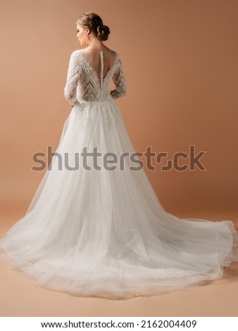Luxury shiny lace wedding dress. Summer backless long sleeve bridal gown with tulle skirt. Beautiful blonde bride posing in studio on brown background. Natural looking makeup and hairstyle, rear view. Royalty-Free Stock Photo #2162004409