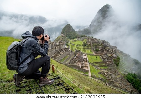 Asian man tourist and photographer taking photo at Machu Picchu, one of seven wonders and famous tourist attraction in Cusco Region of Peru. This majestic place has known as Lost City of the Incas. Royalty-Free Stock Photo #2162005635