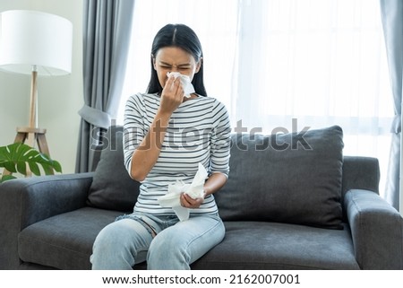 Asian beautiful woman having running nose and sneezing in living room. Attractive young female sitting on sofa feeling bad and suffer from allergy then put tissue cover her nose while sneez in house. Royalty-Free Stock Photo #2162007001