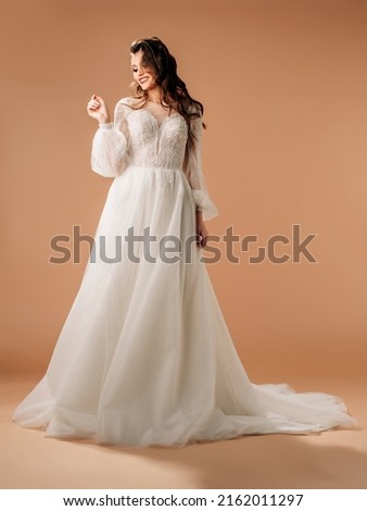 Young brunette bride in elegant white lace wedding dress with long sleeves and lush tulle skirt, big train. Full-length portrait of happy smiling bride in studio. Bridal fashion. Wedding inspiration Royalty-Free Stock Photo #2162011297
