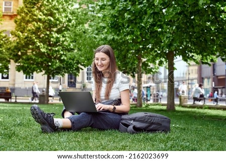 A young girl works sitting on the lawn with a laptop on her lap. A woman takes online courses in the park, sitting on the lawn. The concept of a remote office.