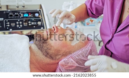  Cosmetologist makes a man a facial therapeutic. Middle-aged man in a spa for beauty treatments. Beautician extrude from  bottle with  moisturizing cream on his hand to apply it to the male face.