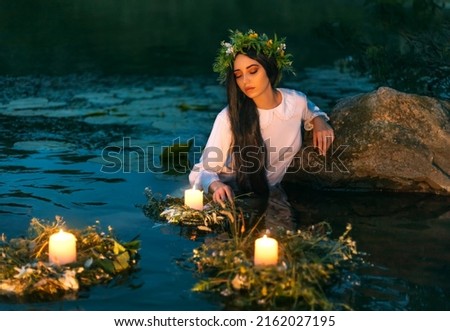 Slavic woman nymph stands in water herbal wreath floa candles burning. Fantasy girl mermaid. White long wet dress. Summer night green grass tree river. pagan holiday Ivan Kupala divination maidens. Royalty-Free Stock Photo #2162027195