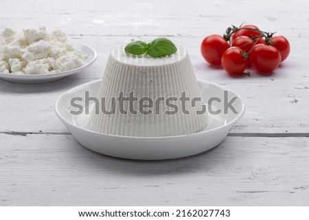 whole cottage cheese with basil leaf resting on white plate with fresh tomatoes resting on white wooden background