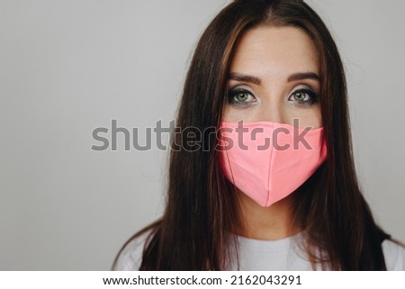 Close-up portrait of girl's face, look straight into camera, her mouth is covered with mask against background of white wall. Girl with make-up, close-up photo without retouching. medicine concept.
